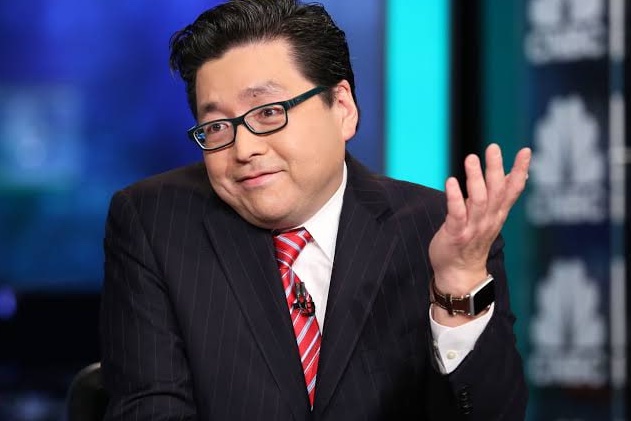 A man in a dark pinstripe suit, red and blue striped tie, and glasses gestures with his right hand raised while appearing to speak on a television set.