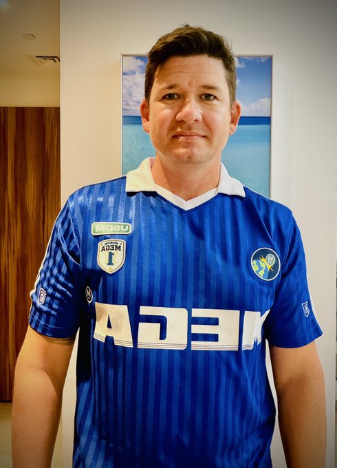 A man posing in a blue soccer jersey featuring 'MEGA' branding and an 'AEL' crest, standing in front of a tropical beach picture.