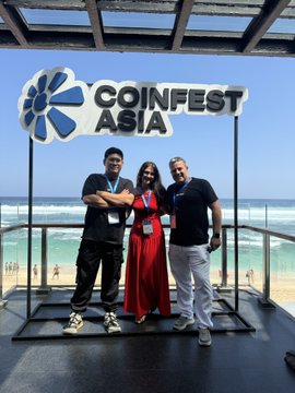 Three people pose in front of a 'COINFEST ASIA' sign at a beachfront location, with the ocean visible in the background, suggesting attendance at the cryptocurrency conference.