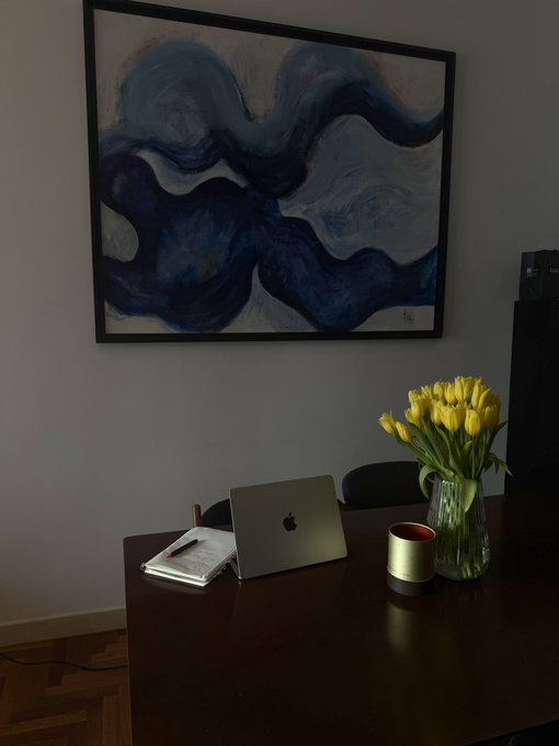 A dark interior scene featuring a wooden table with a laptop, notebook, candle, and vase of yellow tulips, beneath a large framed abstract blue wave painting on a gray wall.