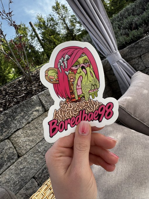 A hand holds up a sticker featuring a stylized illustration of a pink-haired character with the text "Boredhae98" against an outdoor patio background.