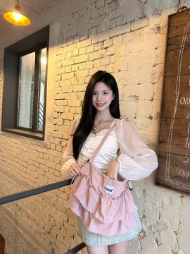 A young woman with long dark hair poses indoors near a brick wall, wearing a white blouse with puffy sleeves, a denim skirt, and carrying a pink frilly handbag.