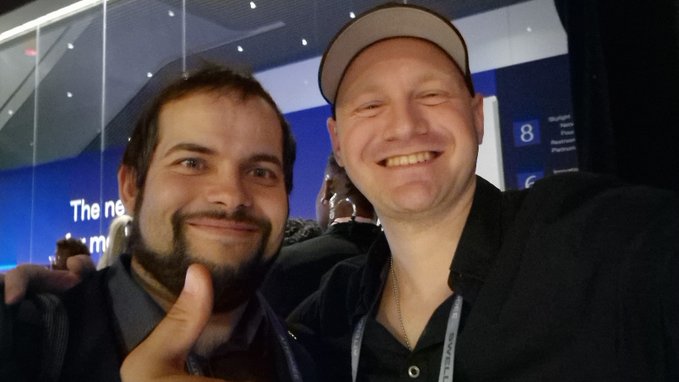 Two men pose for a selfie at an event, with one giving a thumbs-up; a blue sign behind them includes a visible “8” and partial text. The photo accompanies a post about @j00sko explaining borrowing RLUSD against XRP ahead of a planned interview.