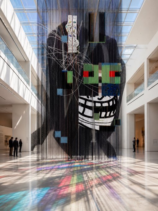 A large suspended art installation depicting a stylized figure hangs in a sunlit atrium, casting colorful pixelated reflections onto the polished floor. The image ties to the post by visually representing the call to "R3ORDR your PFP," with the fragmented, mosaic-like face symbolizing a reinvention or reimagining of one's profile picture.