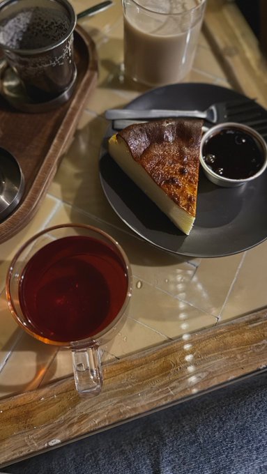An overhead shot of a table setting featuring a slice of burnt Basque cheesecake with a side of berry compote, a glass of tea, and another beverage on a tiled table.