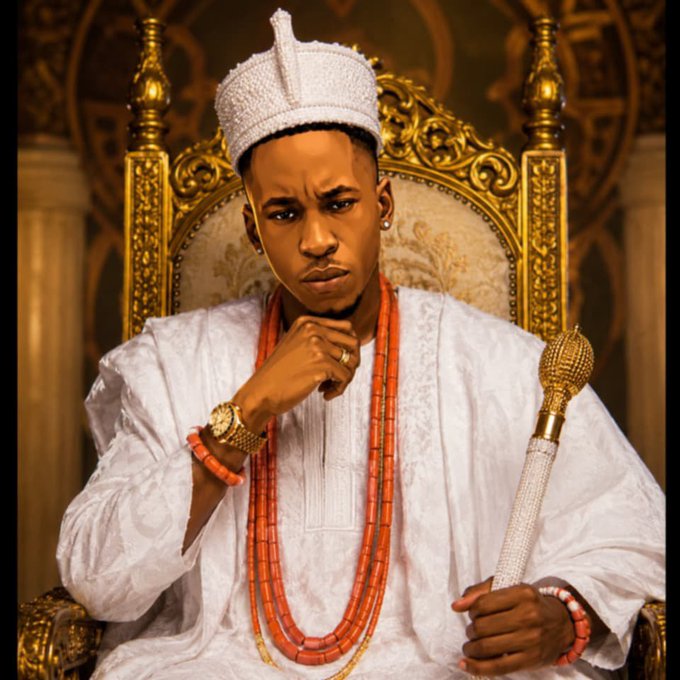 A man wearing a white traditional outfit and coral bead necklaces sits on an ornate golden throne, holding a beaded staff.