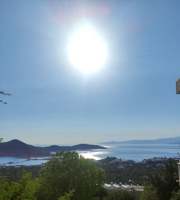 A bright sun shines brilliantly over a serene coastal landscape with distant mountains, shimmering sea, and a small town nestled along the shoreline. The idyllic, tranquil scene contrasts sharply with the bustling online activity mentioned in the post, as the user wakes up to a flood of DMs and vouches from others.