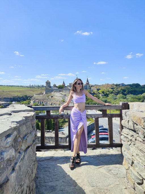 A woman in a lavender two-piece outfit and sunglasses leans against a wooden fence, posing with a historic fortress and green landscape in the background.