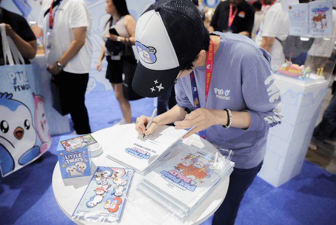 A woman wearing a Pudgy cap and hoodie signs merchandise at a convention booth, with items labeled "Little Thieves" and "Pudgy Penguins" displayed on the table. The scene captures a community member-turned-team member engaging with fans, reflecting the post's celebration of how Pudgy Penguins holders have grown into key roles within the organization.