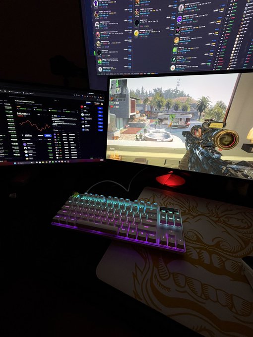 A dimly lit gaming setup features a mechanical keyboard with RGB lighting and two monitors—one displaying a crypto trading chart with a price of "$45,495" and the other showing a first-person shooter game. The setup humorously reinforces the post's joke about being "locked in," as the user has both financial charts and a game open simultaneously, suggesting performative multitasking rather than focused productivity.