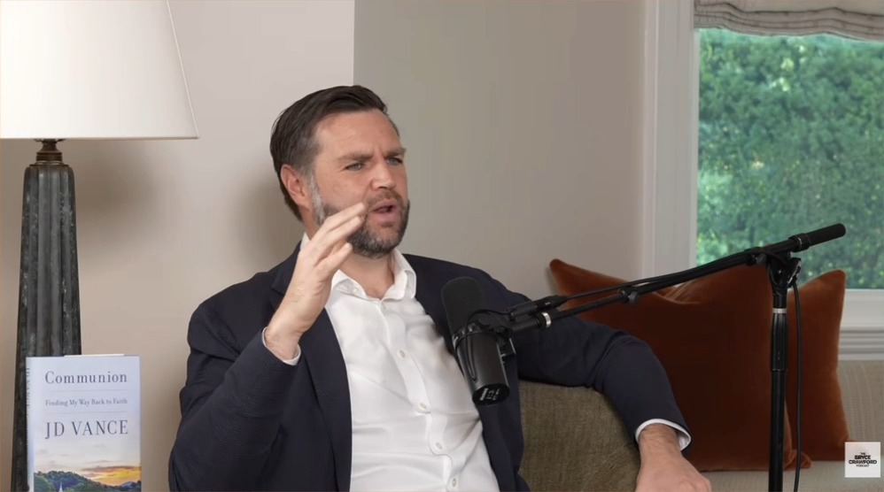 JD Vance, author of 'Communion: Finding My Way Back to Faith,' speaks into a microphone during a podcast interview, with his book displayed beside him.