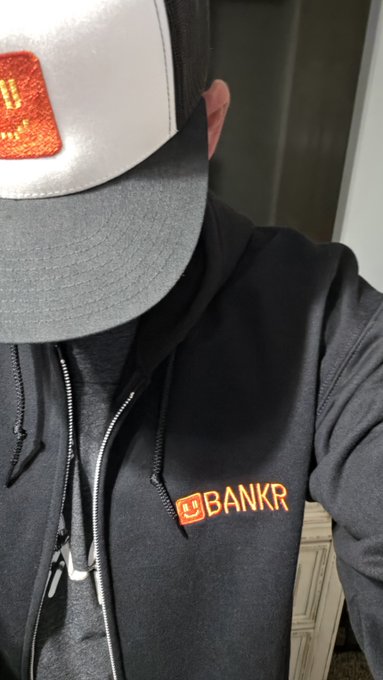 A selfie showing a person wearing a black hoodie with an orange "BANKR" logo, a gray and black trucker hat with an orange patch, and earphones.