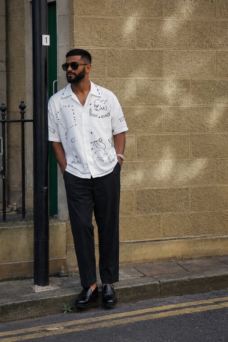 A stylish street fashion photo of a bearded man wearing sunglasses, a white patterned short-sleeve shirt, black trousers, and black loafers, standing on a city street against a stone wall.