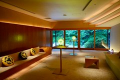 A serene traditional Japanese tea room with tatami mats, built-in wooden seating with cushions, and a view of a lush green garden through large windows.