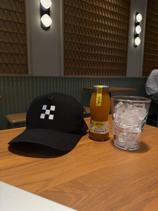 A black cap with a white logo sits on a wooden cafe table next to a bottle of 'multi VITAMIN' drink and a glass of ice water.