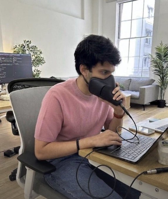 A man at an office desk holds a black desk lamp up to his mouth like a microphone while typing on a keyboard, humorously mimicking someone trying to quietly issue a secret request to CoinStats AI for unannounced meme coins.