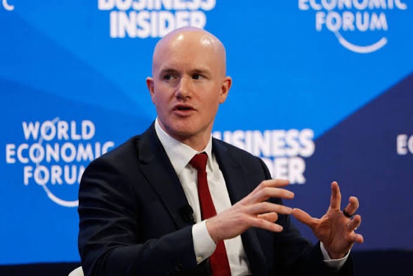 Coinbase CEO Brian Armstrong speaks at the World Economic Forum, gesturing with both hands while wearing a dark suit and red tie, with "Business Insider" and "World Economic Forum" branding visible on the blue backdrop behind him.