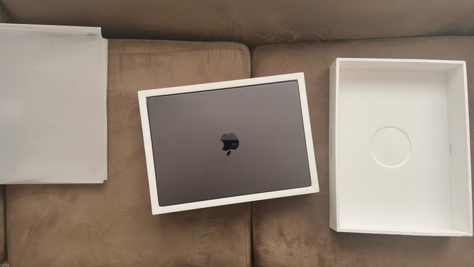 An Apple MacBook in its packaging sits on a brown leather couch, with the white box and lid separated on either side of the laptop.