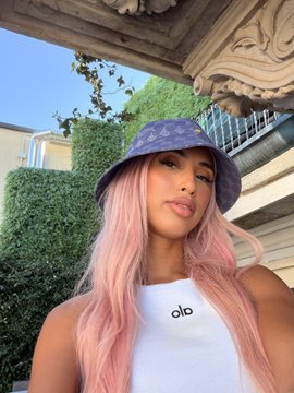 A woman with long pastel pink hair wearing a purple Alo bucket hat and a white Alo tank top poses for a selfie in Milan. The "alo" branding on her clothing aligns with her bullish vibe, matching the caption's positive greeting styled as exclusive access to fellow $ANSEM supporters.