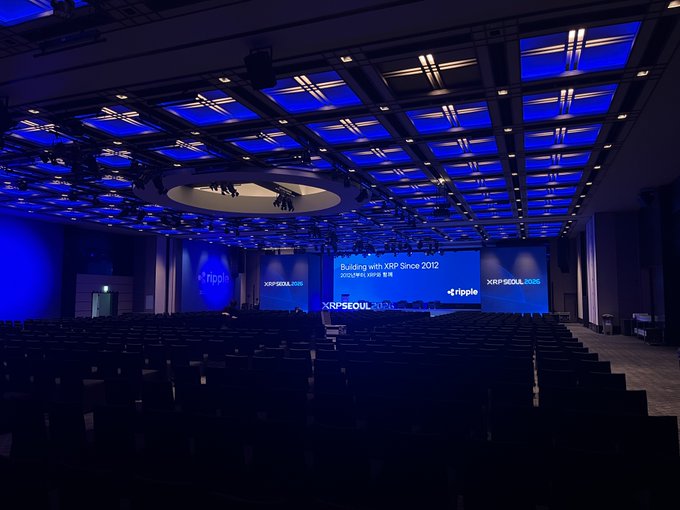 A spacious, empty conference hall illuminated in blue lighting is set up for XRP Seoul 2025, with the main stage screen displaying "Building with XRP Since 2012" alongside the Ripple logo. The image captures the quiet anticipation before the event begins, reflecting Seoul's growing role as a hub for the XRP community.