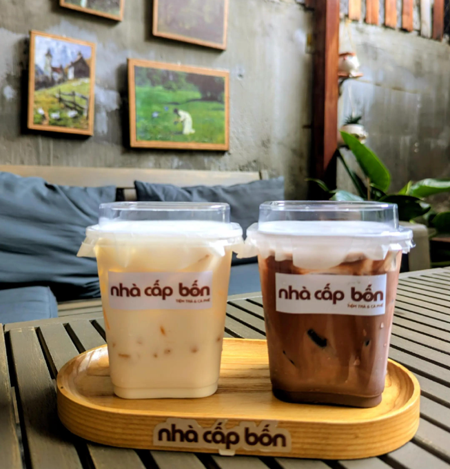 Two iced beverages from "Nhà Cấp Bốn" café sit on a wooden tray with the brand's signature, ready to fuel a fresh week of hustle and good vibes.