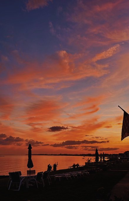 A vertical photograph captures a vibrant, dramatic sunset over a beach or coastal area. The sky is filled with bright orange, red, and deep purple clouds, while silhouettes of lounge chairs, umbrellas, and a distant shoreline are visible below.