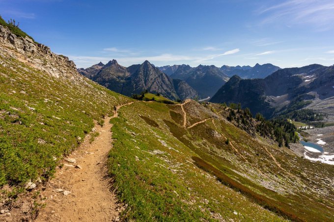 A landscape photograph of a dirt hiking trail winding through grassy alpine terrain, with jagged mountain peaks, scattered pine trees, and patches of snow visible under a blue sky.