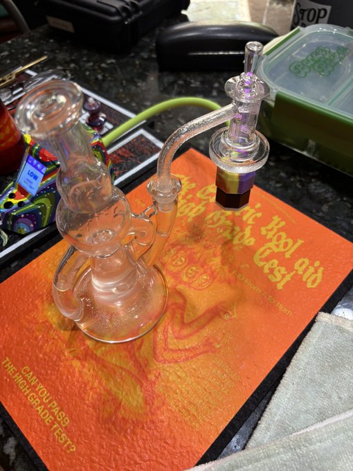 A glass smoking rig (water pipe/dab rig) with an attached recycler attachment and bowl piece sits on an orange mat reading "Don't dab and drive... dab and Cest" and "CAN YOU PASS THE HIGH GRADE TEST?".