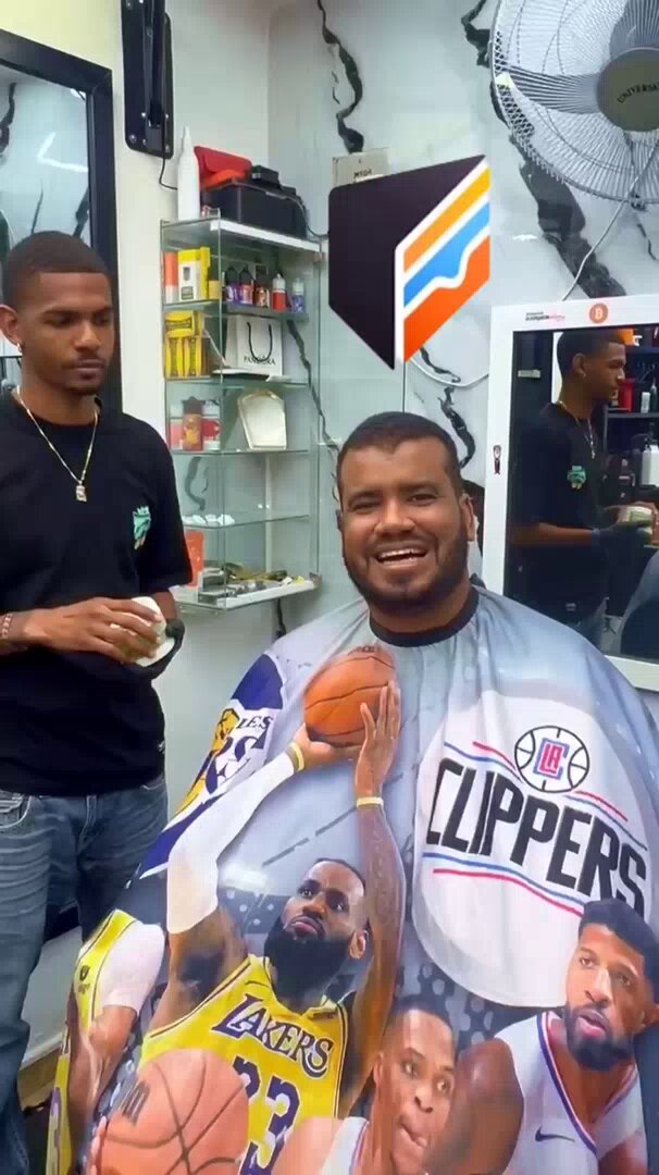 The image features a barber shop with two men inside, one of whom has their head shaved bald and wearing a Giants basketball cap. The other man is smiling at them while they are getting their hair cut. A poster or advertisement for Bitcoin can be seen in the background, indicating that the barbershop may accept Bitcoin as payment.