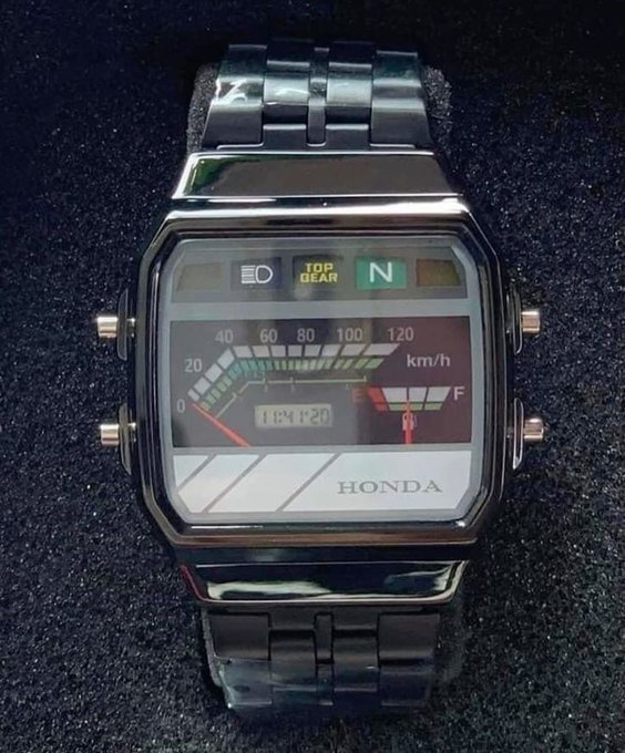 A retro-styled digital watch with a silver metal band, featuring a motorcycle dashboard-inspired display showing speed (km/h), fuel, and the Honda logo, set against a dark textured background.