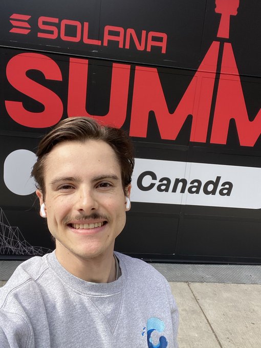 A smiling young man with a mustache takes a selfie wearing white AirPods and a grey sweatshirt, standing in front of a large sign reading 'SOLANA SUMMIT Canada'.