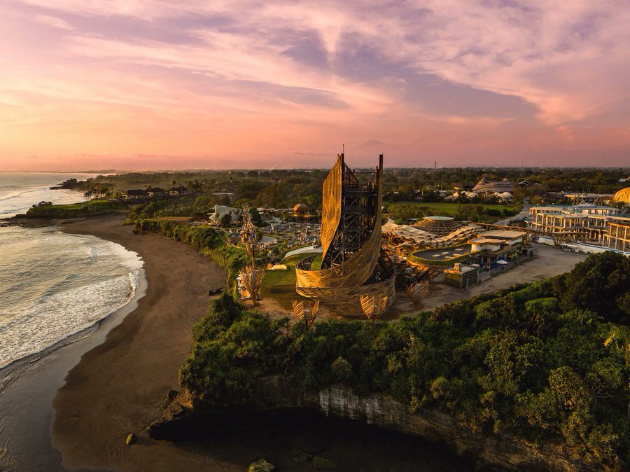 An aerial view of a coastal resort at sunset featuring a distinctive ship-shaped or sail-like building on a cliff overlooking the beach.
