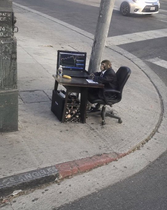A businessman in a suit sits at an outdoor makeshift office on a Los Angeles street corner, intently watching stock charts on a laptop displaying what appears to be GM and CT tickers, embodying the post's "Happy Friday, We go again" sentiment of a trader resuming his market work. A computer tower sits beneath the desk, and "LOS ANGELES" is visible painted on the curb.