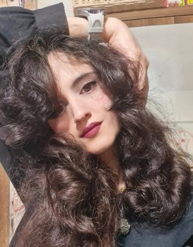 A close-up photo of a person with long, voluminous dark brown curly hair and dark lipstick, posing with their hands raised near their head in an indoor setting.