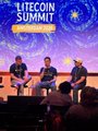 The image features three men sitting behind a podium at an event called "Litecoin Summit" held in America in 2016. The speakers are addressing the audience about the challenges of Bitcoin and its impact on society, with one man speaking directly into the camera. The backdrop is a large screen displaying a starry night sky, adding to the atmosphere of the event.