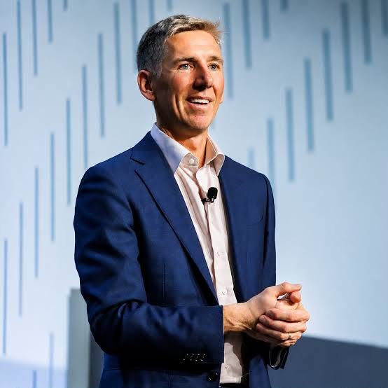 A man in a navy blue suit jacket and light pink shirt standing on a stage with a blue patterned backdrop, smiling while looking upward.