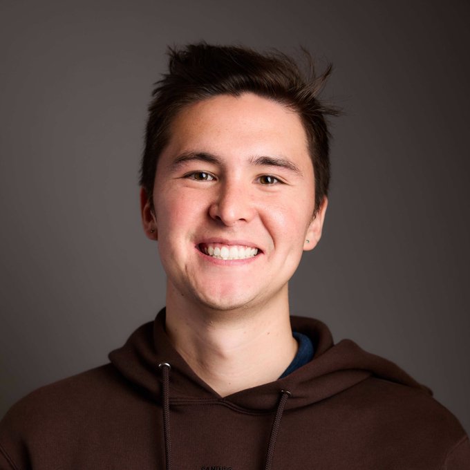 A smiling young man with dark brown hair and a brown hoodie poses for a close-up portrait against a gray backdrop, fitting the caption "New pfp. Who's that guy?" as a fresh profile photo showcasing his friendly, casual look.