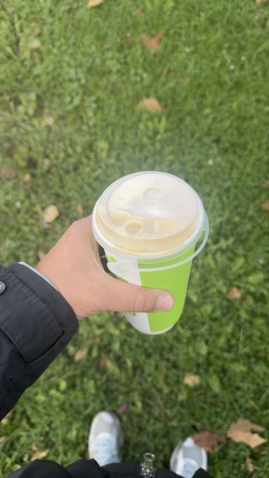 A first-person view of a hand holding a green and white cup of an iced drink over a grassy area with scattered leaves.