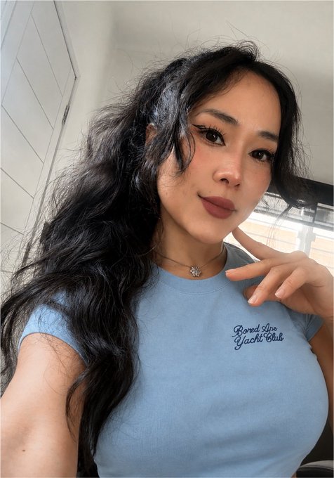 A selfie of a young woman with long dark wavy hair and brown lipstick, wearing a light blue 'Bored Apex Yacht Club' t-shirt and a small pendant necklace.
