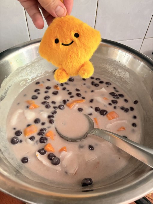 A hand holds a cute yellow plush toy with a smiling face above a metal bowl containing a pink creamy dessert topped with black tapioca pearls, diced fruit, and coconut pieces.