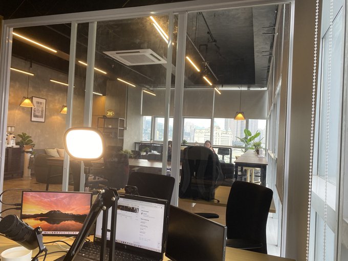 A modern office space with large glass windows features desks with computer monitors, a ring light on a tripod, and a view of the city skyline outside.