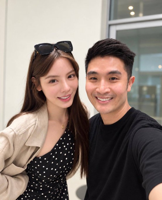 A selfie of a smiling man and woman indoors; the woman has sunglasses perched on her head and wears a polka dot top, while the man wears a black t-shirt.