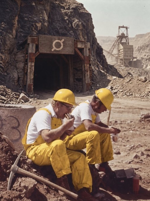 The image shows two men wearing yellow overalls sitting next to each other, with a pile of rocks behind them. They are both eating sandwiches while working at a construction site or a mine. The photo has a vintage filter applied to it, giving it an old-fashioned appearance. The caption "Pivot to mining." suggests that the image is related to a mining project and could be from a time when such projects were more common in the area.