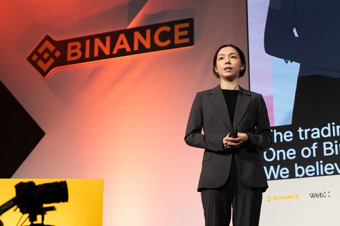A woman in a gray blazer presents on stage in front of a large Binance logo, with a screen displaying partial text about trading and Web3 visible to her right, aligning with the post's discussion of Binance Japan's achievements and market leadership in BTC/JPY and ETH/JPY spot trading.