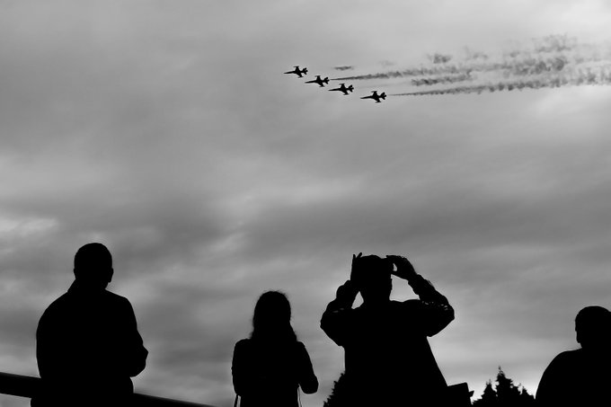 A black and white silhouette photo of spectators watching a formation of jets fly overhead, with one person holding up a phone to take a picture.
