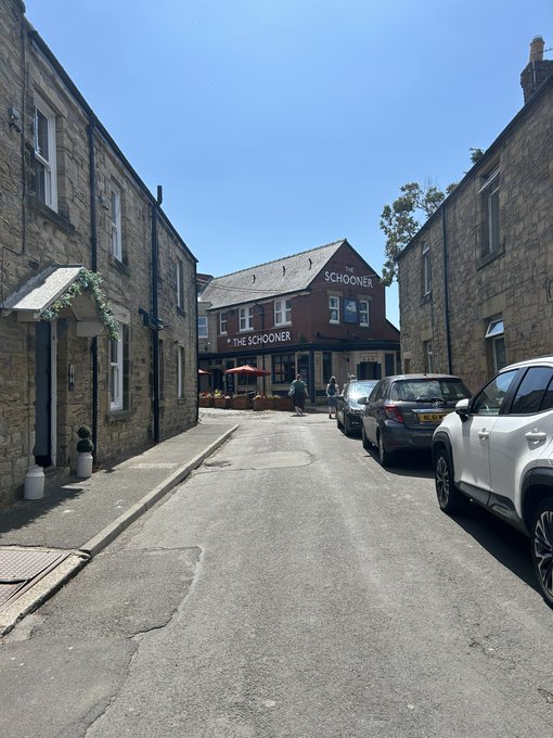 A sunny street view of 'The Schooner' pub, a stone building situated between rows of houses with cars parked along the road.