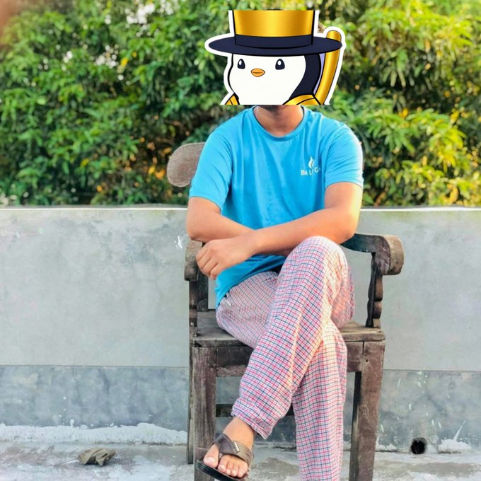 A photo of a person sitting in a wooden chair outdoors wearing a blue t-shirt and checkered pants, with their face obscured by a cartoon penguin sticker wearing a black top hat with a gold band.