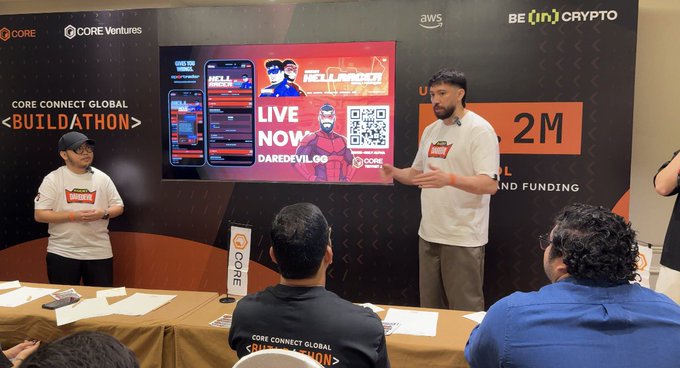 Two men stand on stage presenting at the 'CORE CONNECT GLOBAL <BUILDATHON>' event, with one speaking in front of a screen displaying 'LIVE NOW DAREDEVIL OG' and a QR code.