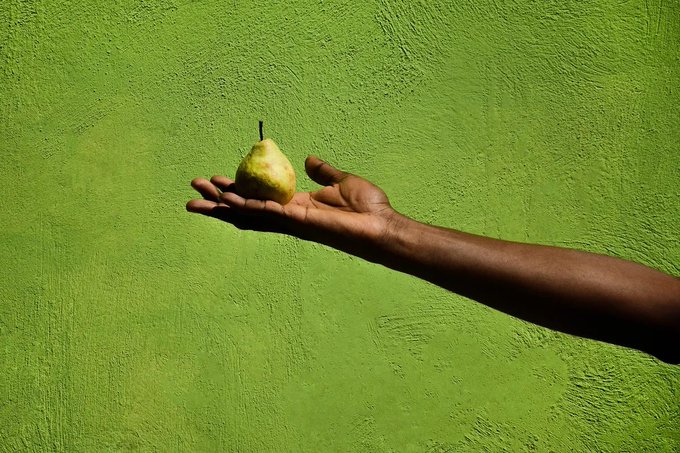 A dark-skinned hand holds a green pear against a textured bright green wall, with strong sunlight casting a distinct shadow.