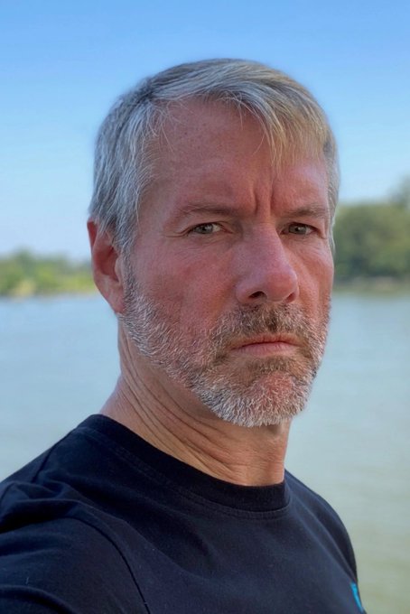 A serious-looking middle-aged man with gray hair and beard stands near a body of water, gazing thoughtfully into the distance. His contemplative expression mirrors the uncertain sentiment of investors evaluating $STRC's modest +3% monthly performance.